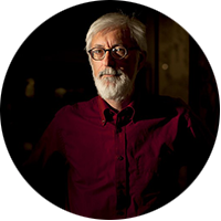 Steven Lubar is a white man with a white hair and beard, wearing glasses and a red shirt, standing in a dark room.