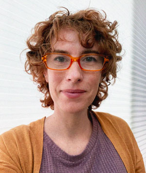 Ginger Nolan, a white woman with short red hair, wears red glasses, gold cardigan and purple top