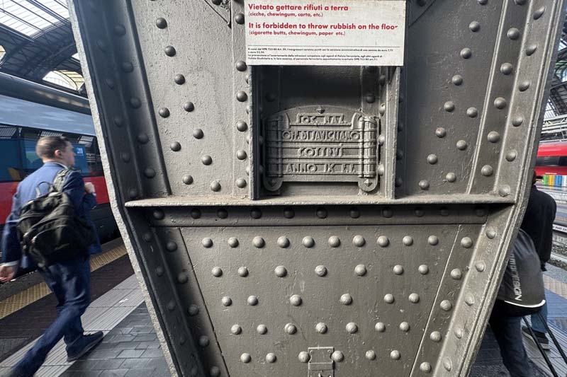 A steel column in the middle of a train platform with people walking by