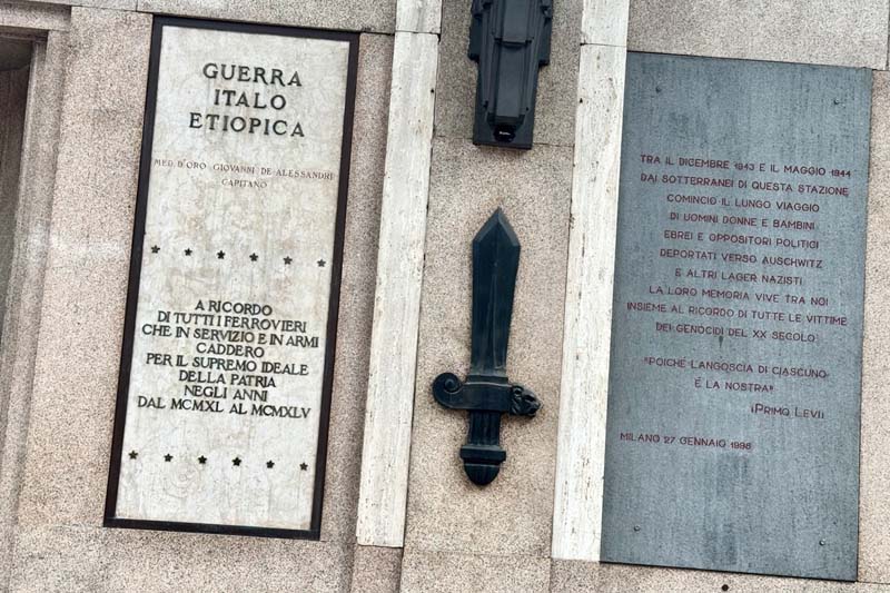 A white sign and a gray sign on a stone wall