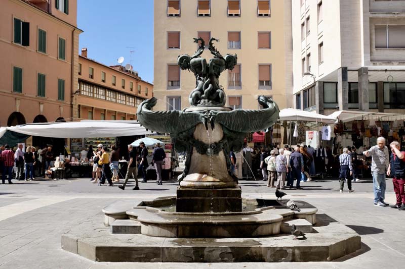 A fountain on a pedestal surrounded by buildings and people walking around