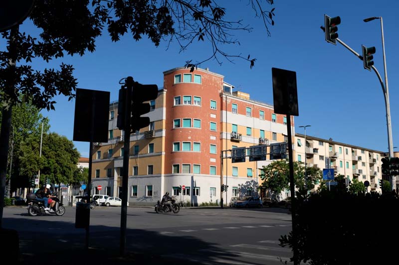 Motorcycles drive by a multi-story orange and yellow building with a rounded corner