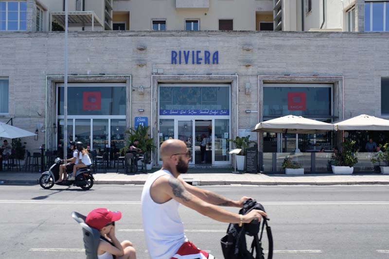 A person with a child biking on a street in front of a building with large windows