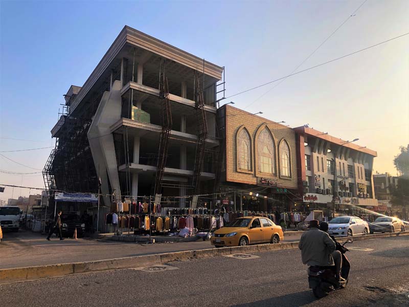 four-story building under construction, clothes hang on racks outside, cars and moped in foreground