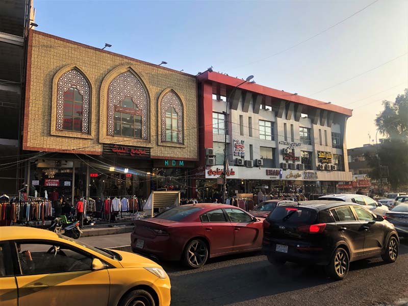 storefronts with racks of clothes on sidewalk, cars on street in foreground