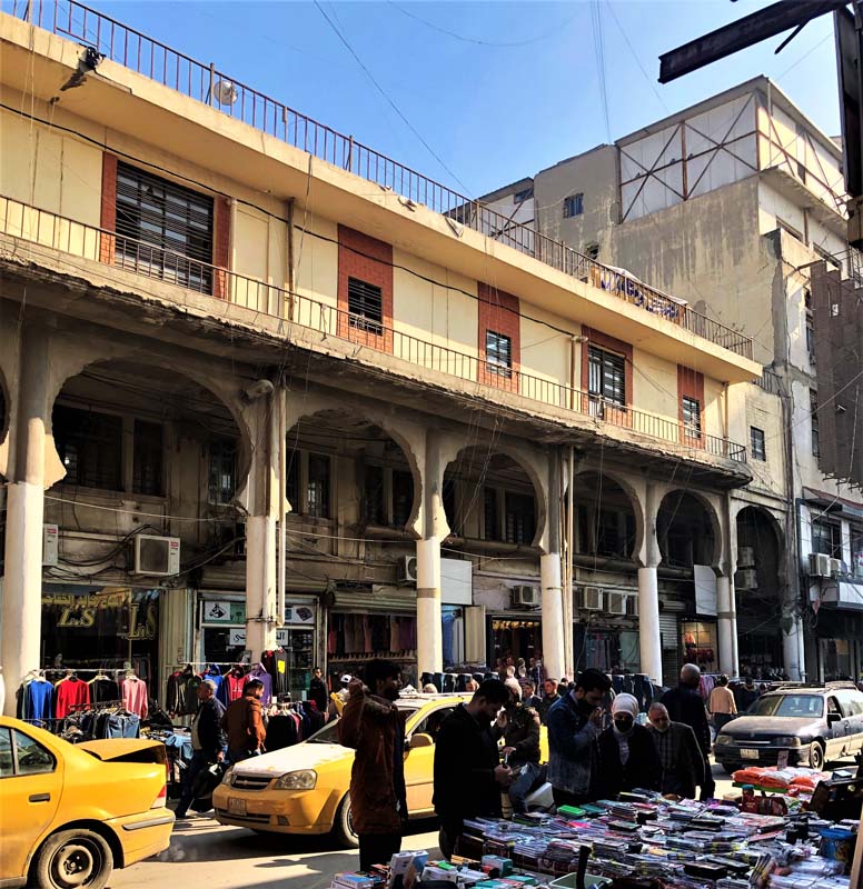 cars are flanked by people and rows of goods, colonnade facade in background