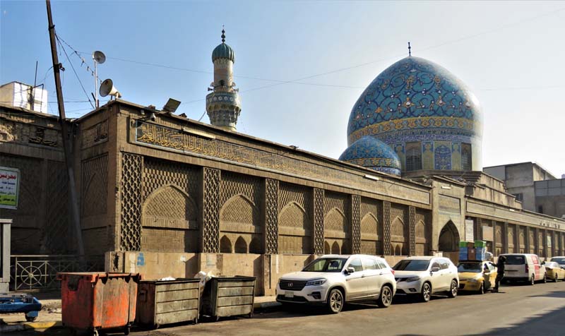 mosque with blue and gold domes, line of parked cars and dumpsters in foreground