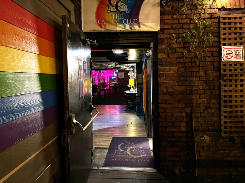 Beside a mural of a rainbow stripes, a metal door in a brick wall is propped open with a doorstop. Beyond, hightop bar tables and colored stage lights invite patrons in. 