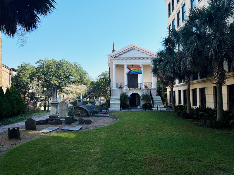  A building with a high-columned porch sits beside a much large building on the right. Weathered tombstones mark the lawn in front.