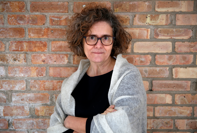 Claire Zimmerman, a white woman with brown hair and glasses, stands against a brick wall