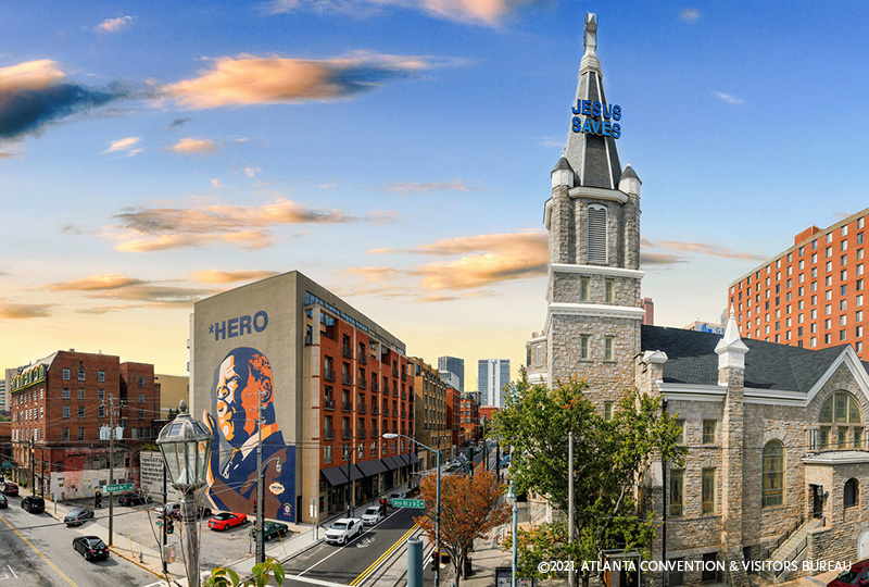 A panoramic sunset view of Atlanta’s Sweet Auburn district, featuring a large mural of a black man labeled “*HERO,” a historic stone church with a tall steeple displaying "JESUS SAVES," and a mix of modern and historic brick buildings lining the streets.