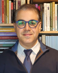 Mohammad Gharipour wears glasses, a white shirt and blue tie, a black jacket, in front of a bookcase