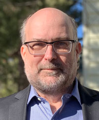 Eric Mumford, a bald man with a gray goatee, wears glasses, a blue shirt, and a gray jacket