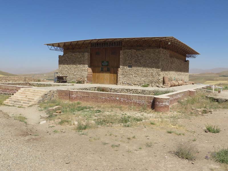 A short flight of stairs leads to a small masonry building with large wooden doors and a flat roof standing in a desert landscape