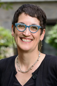 Abigail Van Slyck, a white woman with cropped brown hair, wears blue glasses, long earrings, a necklace, and a black shirt and cardigan.