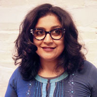 Anooradha Siddiqi, a woman with long brown hair, wears black glasses and a blue shirt