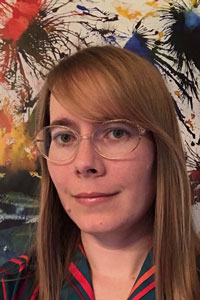 Ashley Gardini, a woman with long blond hair and glasses, wears a black and red striped shirt against a colorful background of paint splashes