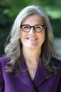Dianne Harris, a white woman with long, gray hair, wears glasses, a purple blazer, and smiles at the camera