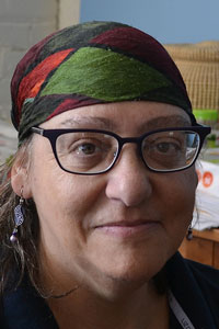 Gail Dubrow, a white woman with shoulder-length hair, faces the camera and wears a patterned scarf, black glasses, and earrings.
