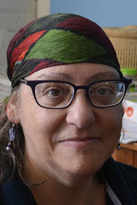 Gail Dubrow, a white woman with shoulder-length hair, faces the camera and wears a patterned scarf, black glasses, and earrings.