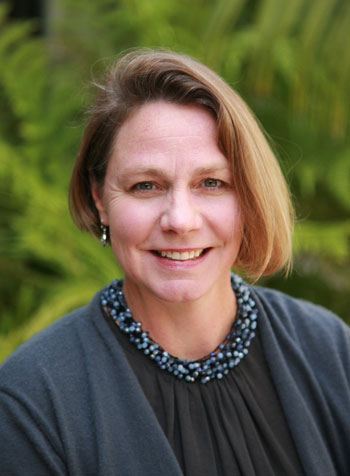 Bridget Maley, a white woman with short blond hair, wears a navy top and cardigan, blue necklace, and smiles at the camera