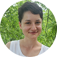 Perri, a white woman with short brown hair, smiles at the camera and wears a sleeveless white blouse. She leans against a wooden deck with foliage in the background.