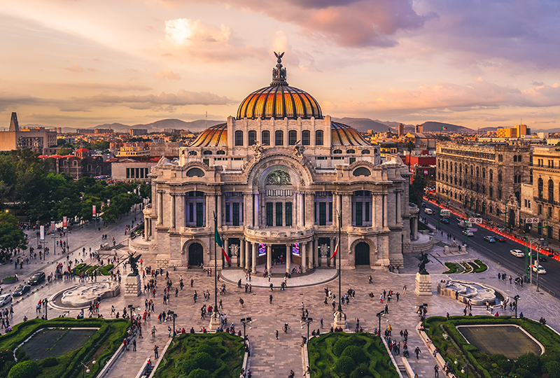 The image shows the Palacio de Bellas Artes in Mexico City, with its grand dome and neoclassical architecture. People walk around the plaza in front, surrounded by greenery. The sunset sky casts a warm glow over the building and the distant cityscape.
