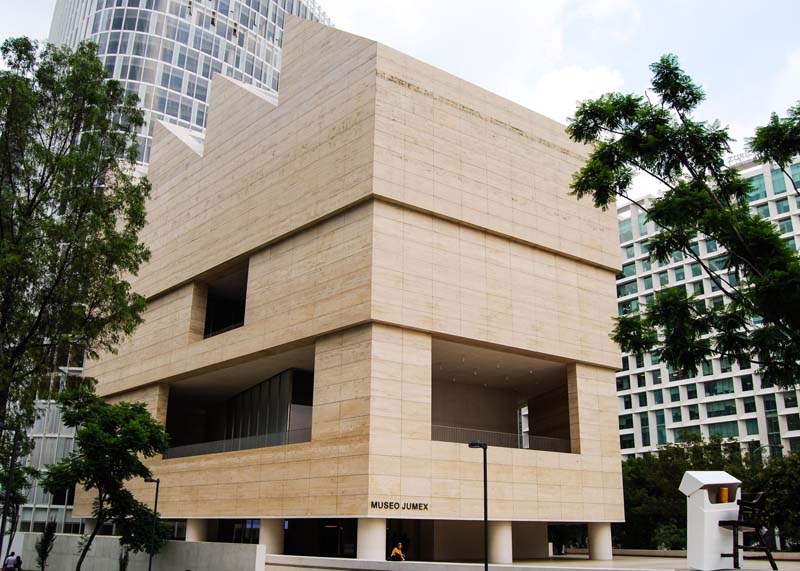 A tan modular building surrounded by trees sand taller buildings