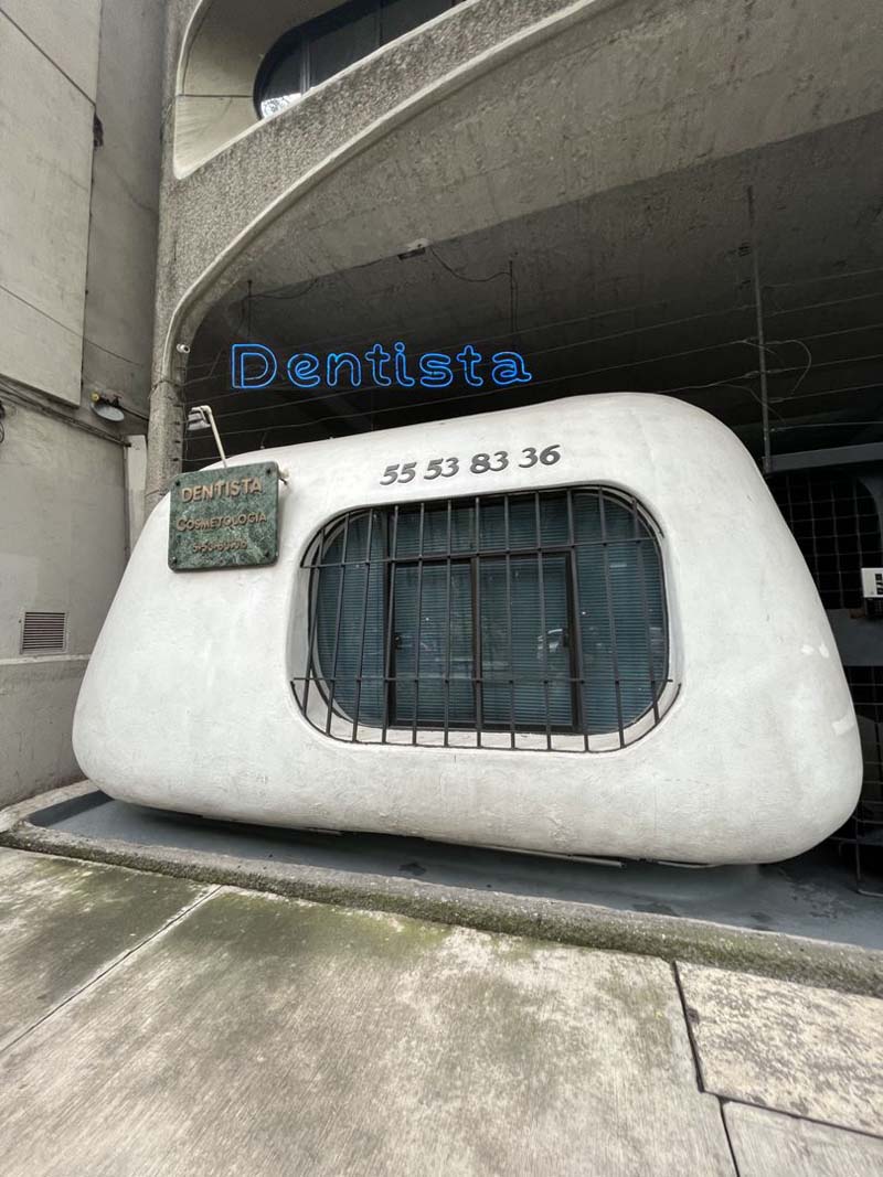 A small, white tooth-shaped building with a window in front of a taller gray building