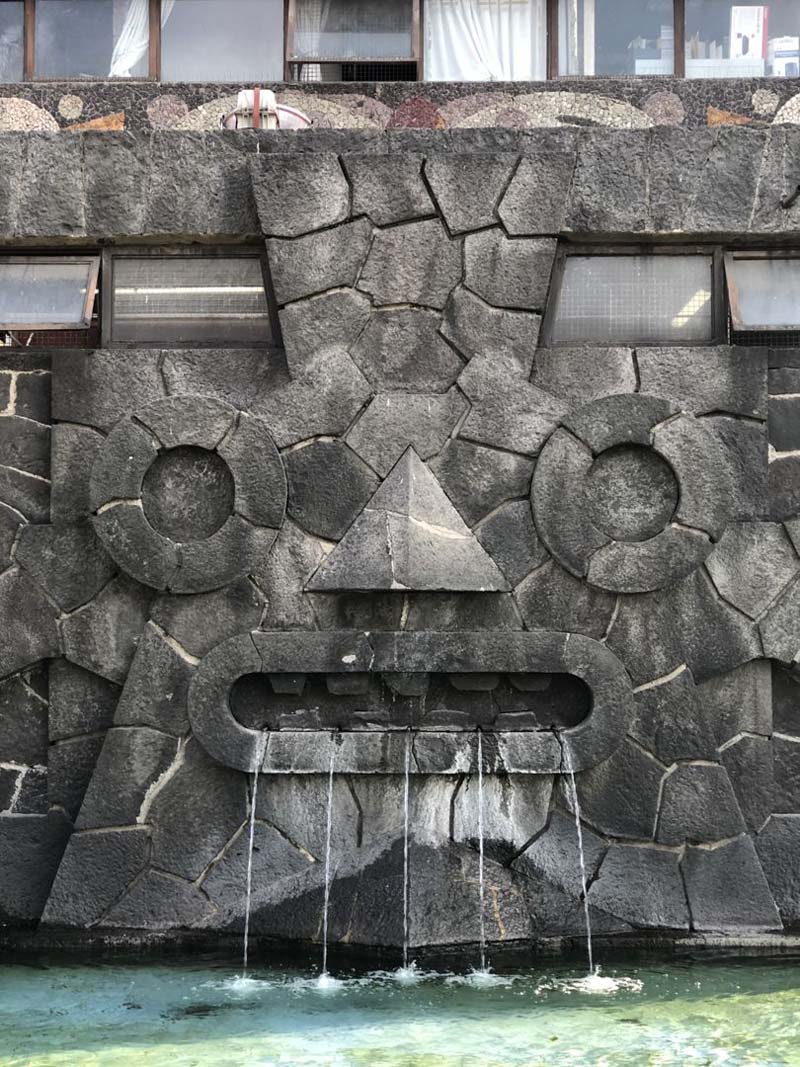 A stone facade resembling a face with geometric shapes forming the eyes, nose, and mouth. Water flows from the mouth into a pool below. The structure is part of a larger building with windows visible at the top.