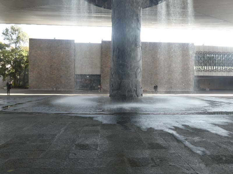 A large stone pillar stands in the center of an open courtyard, supporting a wide roof. Water cascades down around the pillar, creating a curtain of water that splashes onto the ground below. A building stands in the background, with a few people walking or standing in front.