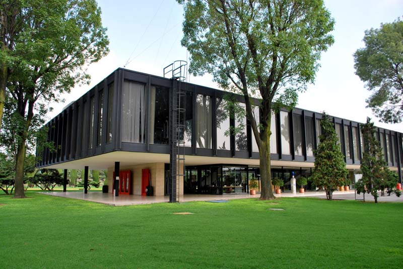 A two-story building with a black exterior and large glass windows, surrounded by green grass and tall trees. The building has an open ground floor with visible entrances and red accents.