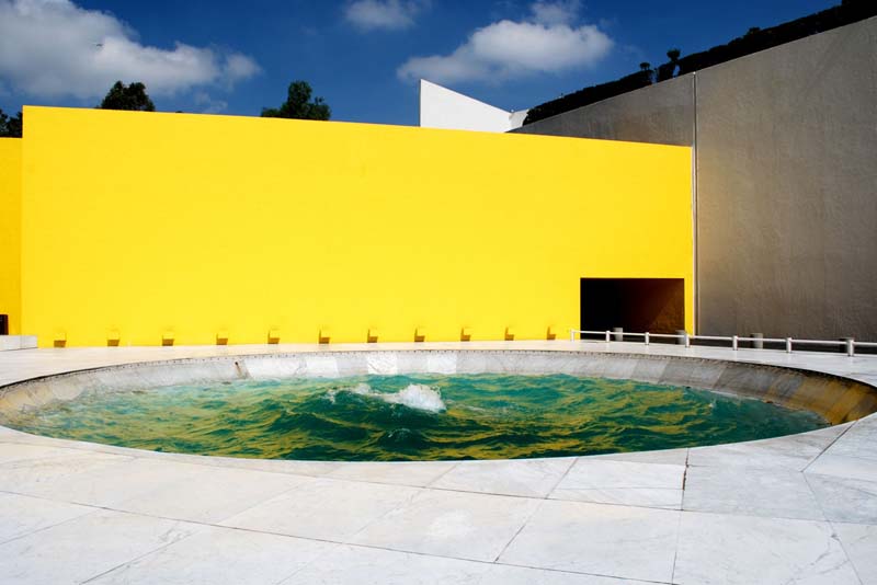 A circular water feature is set against a bright yellow wall. The sky is blue with scattered clouds, and trees are visible in the background.