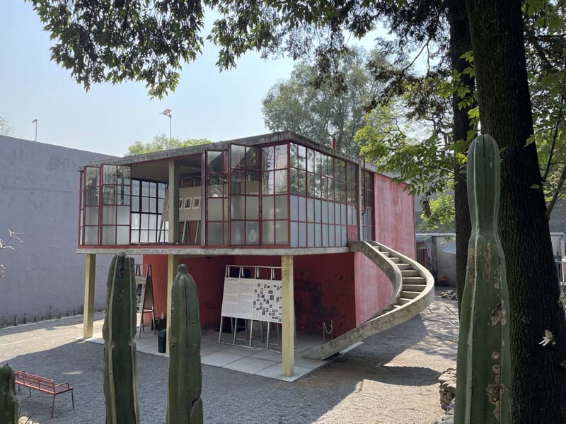A two-story building with a red exterior and large glass windows. A curved staircase connects the ground to the upper level. Tall cacti and surrounding trees add greenery to the scene.