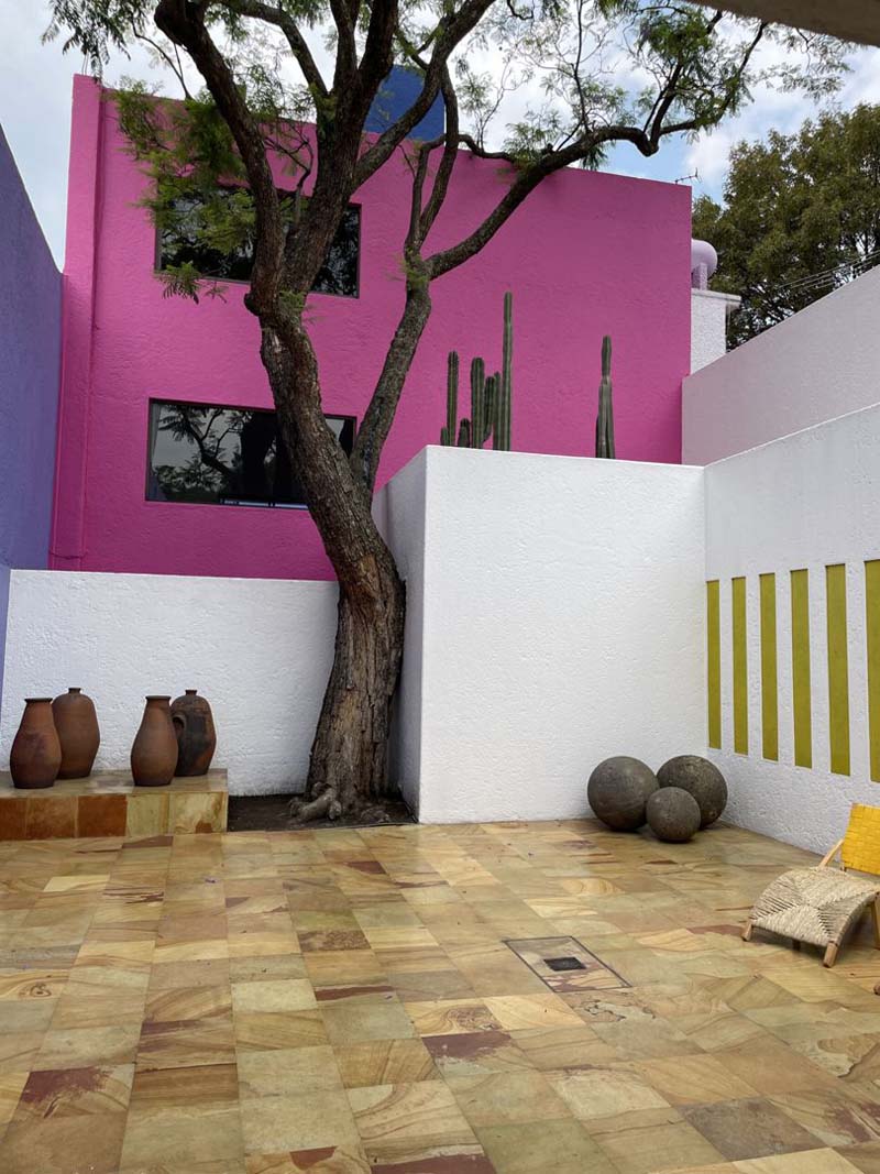 A bright pink building stands behind a courtyard with brown tiles and a tall tree