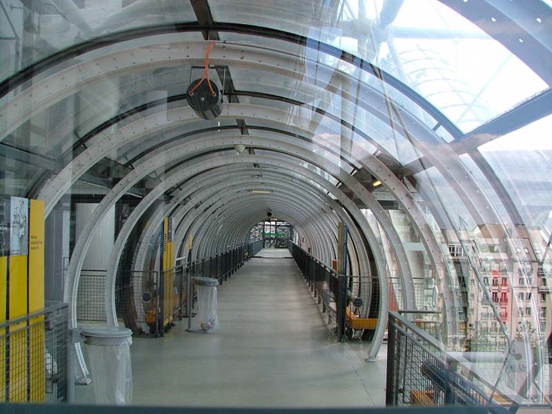 steel and glass arched walkway with concrete floors, yellow panels, metal grate railings