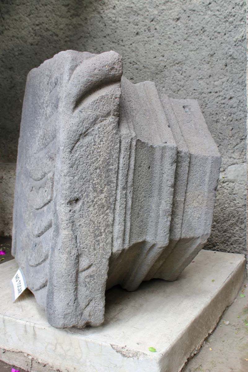 A large, weathered stone artifact displayed on a concrete pedestal. The stone has a rough texture and detailed patterns along its edges. A small white label is placed near the base, and the background features a textured gray wall