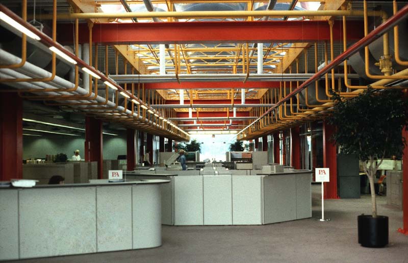 office space with cubicles along center, red and yellow steel ceiling beams with pipes, concrete floor