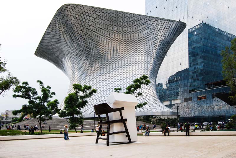 A large,  silver-colored abstract building with hexagon shapes sits next to a taller building with glass windows. 